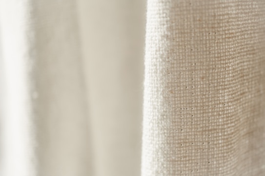 A close-up view of a textured fabric with a light cream color. The weave pattern of the textile is visible, highlighting the natural fibers and intricate details.