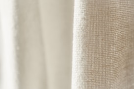 A close-up view of a textured fabric with a light cream color. The weave pattern of the textile is visible, highlighting the natural fibers and intricate details.