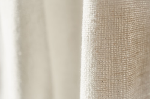 A close-up view of a textured fabric with a light cream color. The weave pattern of the textile is visible, highlighting the natural fibers and intricate details.