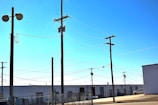 Multiple utility poles with interconnected power lines span across a fenced industrial area. The background includes warehouses with roll-up doors, and a parked white truck is visible. The ground is paved, and the scene is under a clear, blue sky.