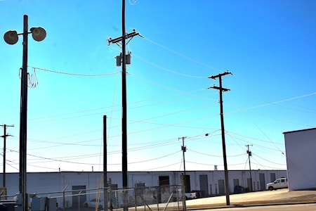 Multiple utility poles with interconnected power lines span across a fenced industrial area. The background includes warehouses with roll-up doors, and a parked white truck is visible. The ground is paved, and the scene is under a clear, blue sky.