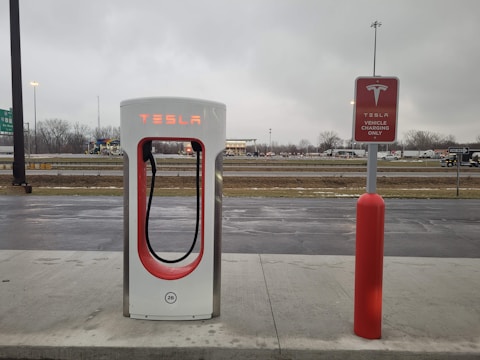 An electric vehicle charging station with the Tesla logo is situated at a roadside area. The charging unit is white with red accents and is positioned on a concrete platform. In the background, there is a highway with vehicles passing by, and the sky is overcast, suggesting a cloudy day.