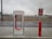 An electric vehicle charging station with the Tesla logo is situated at a roadside area. The charging unit is white with red accents and is positioned on a concrete platform. In the background, there is a highway with vehicles passing by, and the sky is overcast, suggesting a cloudy day.