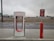 An electric vehicle charging station with the Tesla logo is situated at a roadside area. The charging unit is white with red accents and is positioned on a concrete platform. In the background, there is a highway with vehicles passing by, and the sky is overcast, suggesting a cloudy day.