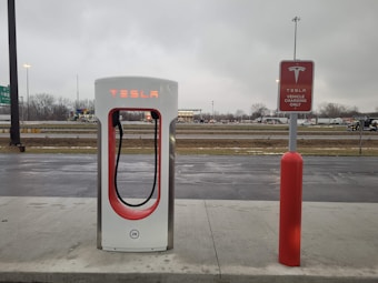 An electric vehicle charging station with the Tesla logo is situated at a roadside area. The charging unit is white with red accents and is positioned on a concrete platform. In the background, there is a highway with vehicles passing by, and the sky is overcast, suggesting a cloudy day.