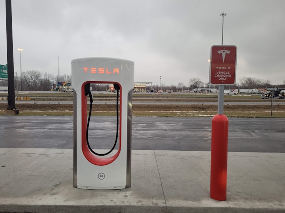 An electric vehicle charging station with the Tesla logo is situated at a roadside area. The charging unit is white with red accents and is positioned on a concrete platform. In the background, there is a highway with vehicles passing by, and the sky is overcast, suggesting a cloudy day.