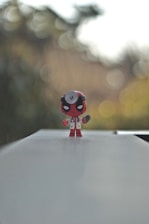 A small figurine with a red and black color scheme stands prominently on a flat, light-colored surface. The character is dressed in a doctor's coat and has a stethoscope. The background is blurred with bokeh effects, creating a serene and focused scene.