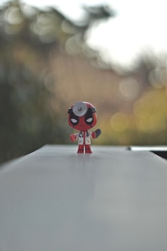 A small figurine with a red and black color scheme stands prominently on a flat, light-colored surface. The character is dressed in a doctor's coat and has a stethoscope. The background is blurred with bokeh effects, creating a serene and focused scene.