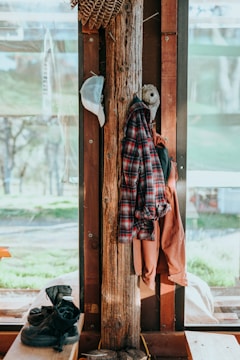 A wooden post indoors is used as a makeshift hanger. A plaid shirt and a jacket are draped over it, with a hat hanging from a nail. At the bottom right, a pair of black boots sit on a bench. The scene is lit with natural light from large windows, offering a view of a lush, green outdoor environment.
