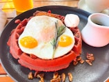 A vibrant red waffle topped with a sunny side up fried egg, garnished with rosemary, pecans scattered around the plate, and accompanied by a small cup of syrup and a dollop of cream.