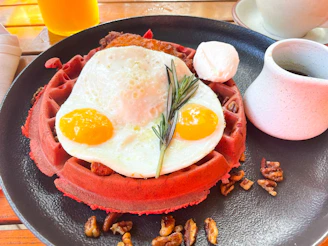 A vibrant red waffle topped with a sunny side up fried egg, garnished with rosemary, pecans scattered around the plate, and accompanied by a small cup of syrup and a dollop of cream.