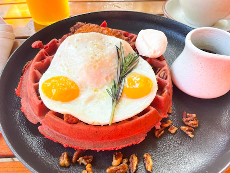 A vibrant red waffle topped with a sunny side up fried egg, garnished with rosemary, pecans scattered around the plate, and accompanied by a small cup of syrup and a dollop of cream.
