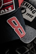 A close-up view of two baseball caps with detailed embroidery. The foreground cap features the number '8' in bold red embroidery on a dark fabric. Another cap in the background has the word '1992' stitched on its patch, alongside a partially visible graphic.