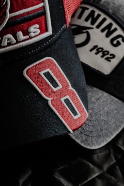 A close-up view of two baseball caps with detailed embroidery. The foreground cap features the number '8' in bold red embroidery on a dark fabric. Another cap in the background has the word '1992' stitched on its patch, alongside a partially visible graphic.
