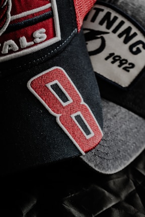 A close-up view of two baseball caps with detailed embroidery. The foreground cap features the number '8' in bold red embroidery on a dark fabric. Another cap in the background has the word '1992' stitched on its patch, alongside a partially visible graphic.