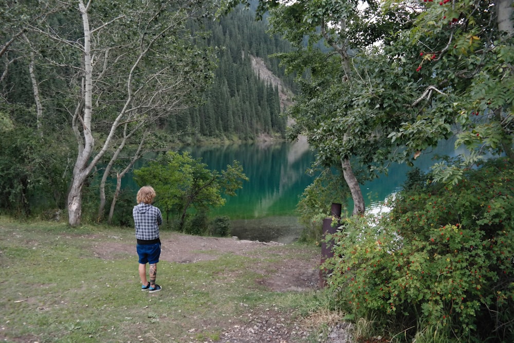 a young boy standing in front of a lake surrounded by trees