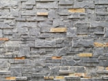 A textured stone wall composed of irregularly shaped gray and brown stones, arranged neatly in a layered pattern. The stones vary in size and have a rough surface, showcasing natural lines and veins.