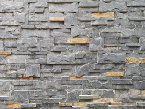 A textured stone wall composed of irregularly shaped gray and brown stones, arranged neatly in a layered pattern. The stones vary in size and have a rough surface, showcasing natural lines and veins.