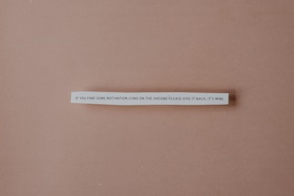 A strip of paper with a humorous quote is centered against a plain brown background. The text reads: 'If you find some motivation lying on the ground please give it back, it's mine.'