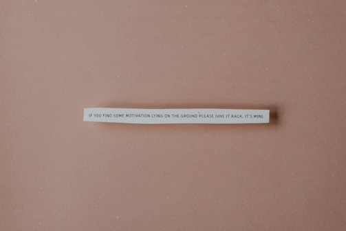 A strip of paper with a humorous quote is centered against a plain brown background. The text reads: 'If you find some motivation lying on the ground please give it back, it's mine.'