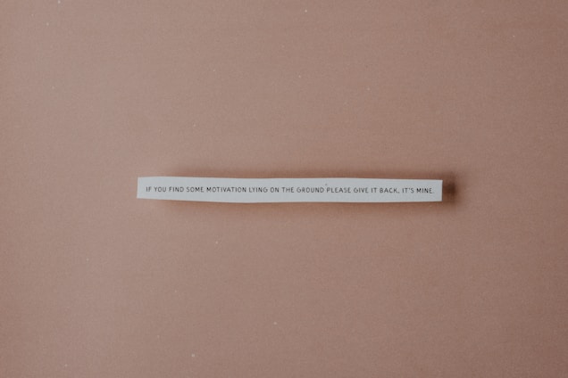 A strip of paper with a humorous quote is centered against a plain brown background. The text reads: 'If you find some motivation lying on the ground please give it back, it's mine.'