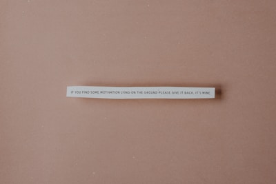 A strip of paper with a humorous quote is centered against a plain brown background. The text reads: 'If you find some motivation lying on the ground please give it back, it's mine.'