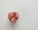 Three pink roses arranged in a heart-shaped white vase are positioned against a white, textured background. The roses have delicate, layered petals with hints of deeper pink at the edges.