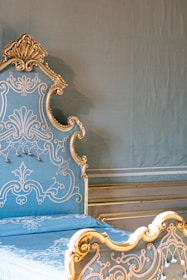 An ornate bed with intricate gold detailing against a muted green wall. The headboard and footboard feature elaborate scrollwork and floral patterns in gold, contrasted against a light blue upholstered fabric. The setting suggests a luxurious and vintage interior design.
