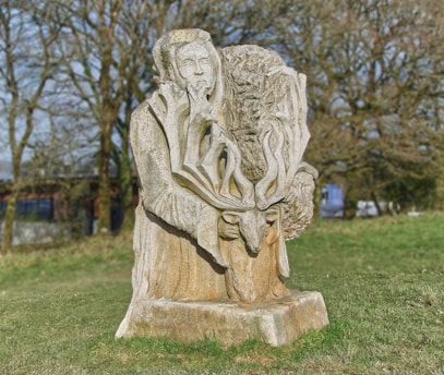 A stone sculpture standing in a grassy outdoor area, featuring a figure with a beard and intricate detailing around the hands, surrounded by tree and branch motifs. The sculpture seems to merge human and natural forms.