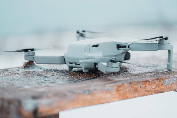 A compact, modern drone is resting on a wooden surface dusted with snow. The drone is gray and streamlined, with extended rotor arms and visible camera components. The background is softly blurred, suggesting a cold outdoor environment.
