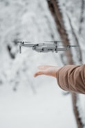 A drone hovers near an outstretched hand against a blurred snowy background. The person is wearing a brown coat, and the scene conveys a sense of interaction between technology and nature.