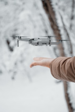 A drone hovers near an outstretched hand against a blurred snowy background. The person is wearing a brown coat, and the scene conveys a sense of interaction between technology and nature.
