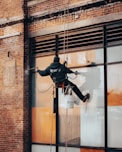 A worker is suspended on ropes, cleaning the windows of a large brick building. The person is equipped with cleaning gear, including a brush and a safety harness attached to a bucket of supplies. The window panes have visible streaks being cleaned, contrasted against the industrial background of brick and metal.