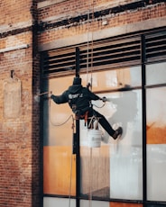 A worker is suspended on ropes, cleaning the windows of a large brick building. The person is equipped with cleaning gear, including a brush and a safety harness attached to a bucket of supplies. The window panes have visible streaks being cleaned, contrasted against the industrial background of brick and metal.