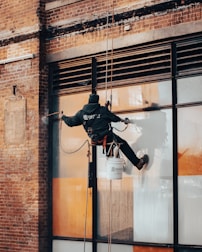 A worker is suspended on ropes, cleaning the windows of a large brick building. The person is equipped with cleaning gear, including a brush and a safety harness attached to a bucket of supplies. The window panes have visible streaks being cleaned, contrasted against the industrial background of brick and metal.