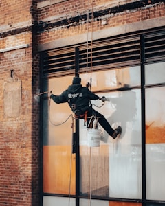 A worker is suspended on ropes, cleaning the windows of a large brick building. The person is equipped with cleaning gear, including a brush and a safety harness attached to a bucket of supplies. The window panes have visible streaks being cleaned, contrasted against the industrial background of brick and metal.