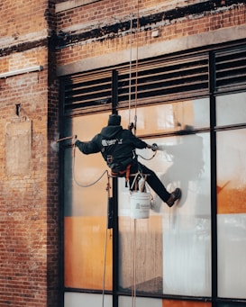 A worker is suspended on ropes, cleaning the windows of a large brick building. The person is equipped with cleaning gear, including a brush and a safety harness attached to a bucket of supplies. The window panes have visible streaks being cleaned, contrasted against the industrial background of brick and metal.