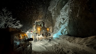 A snow-covered road during nighttime with a snowplow clearing the path. Snow is falling heavily, and the ground and surrounding trees are blanketed in white. The headlights of the snowplow and a parked vehicle illuminate the scene, casting shadows on the rocky cliffside.