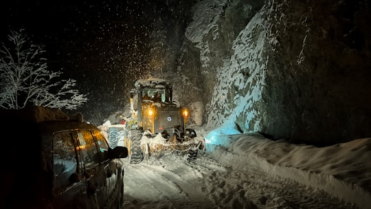 A snow-covered road during nighttime with a snowplow clearing the path. Snow is falling heavily, and the ground and surrounding trees are blanketed in white. The headlights of the snowplow and a parked vehicle illuminate the scene, casting shadows on the rocky cliffside.