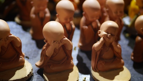 Several small, clay figurines of monks are arranged in a group. Each monk has a joyful expression, and their hands are positioned in playful gestures, creating a whimsical and serene atmosphere. The lighting highlights the smooth texture of the clay and casts gentle shadows.