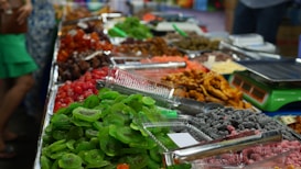 A colorful assortment of dried fruits and snacks is displayed on a market stall. There are trays filled with vibrant green kiwi slices, bright red cherries, and other assorted dried fruits in various shades of red, orange, and brown. Alongside the trays are some electronic scales, suggesting this is a place where items are sold by weight. The setup is neatly arranged, and the fruits have a glossy appearance.