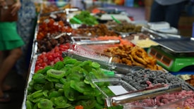 A colorful assortment of dried fruits and snacks is displayed on a market stall. There are trays filled with vibrant green kiwi slices, bright red cherries, and other assorted dried fruits in various shades of red, orange, and brown. Alongside the trays are some electronic scales, suggesting this is a place where items are sold by weight. The setup is neatly arranged, and the fruits have a glossy appearance.
