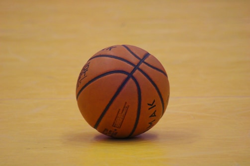A single basketball rests on a hardwood floor, with visible seams and branding on the ball's surface. The floor's light wood tone contrasts with the orange of the basketball.
