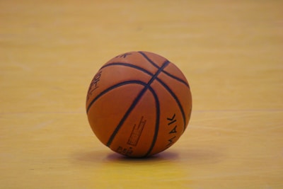 A single basketball rests on a hardwood floor, with visible seams and branding on the ball's surface. The floor's light wood tone contrasts with the orange of the basketball.