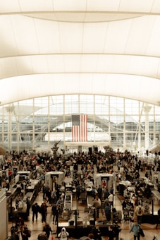 A busy airport security checkpoint area with many people in line, passing through metal detectors. The space is large and spacious, with an American flag hanging prominently from the ceiling. The area is filled with natural light streaming through large windows.