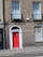 A brick building facade featuring a vibrant red door framed with white trim and pillars. Above the door, there is a semi-circular window with an arch. The building has two large windows with black wrought iron railings. The outer walls are covered in traditional brickwork, contributing to a classic architectural style.