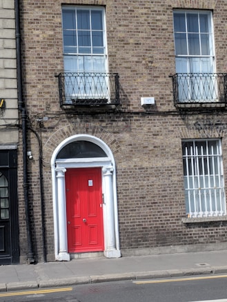 A brick building facade featuring a vibrant red door framed with white trim and pillars. Above the door, there is a semi-circular window with an arch. The building has two large windows with black wrought iron railings. The outer walls are covered in traditional brickwork, contributing to a classic architectural style.