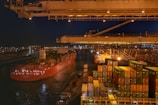 A bustling port scene at night featuring a large cargo ship loaded with colorful shipping containers. The ship is docked beside a terminal with stacked containers and large cranes overhead, while city lights illuminate the background.