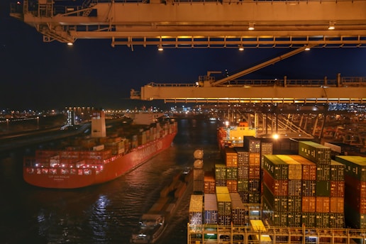 A bustling port scene at night featuring a large cargo ship loaded with colorful shipping containers. The ship is docked beside a terminal with stacked containers and large cranes overhead, while city lights illuminate the background.