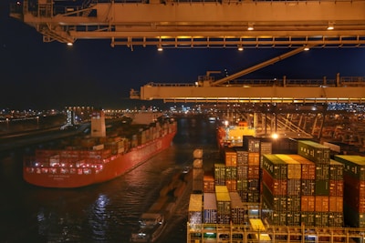 A bustling port scene at night featuring a large cargo ship loaded with colorful shipping containers. The ship is docked beside a terminal with stacked containers and large cranes overhead, while city lights illuminate the background.